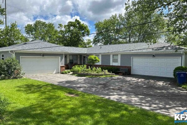 Ranch-style home with light gray vinyl siding, white garage doors, brick foundation accent, and covered front porch